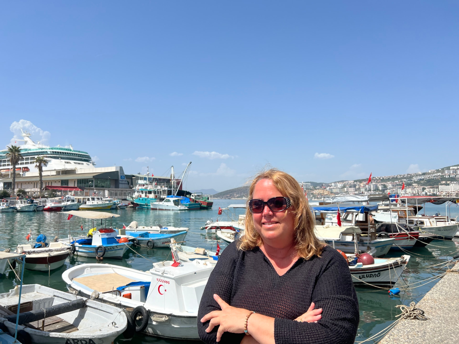 Christine Scheeler standing along the harbor in Kusadasi, Turkey, with fishing boats and a cruise ship behind her