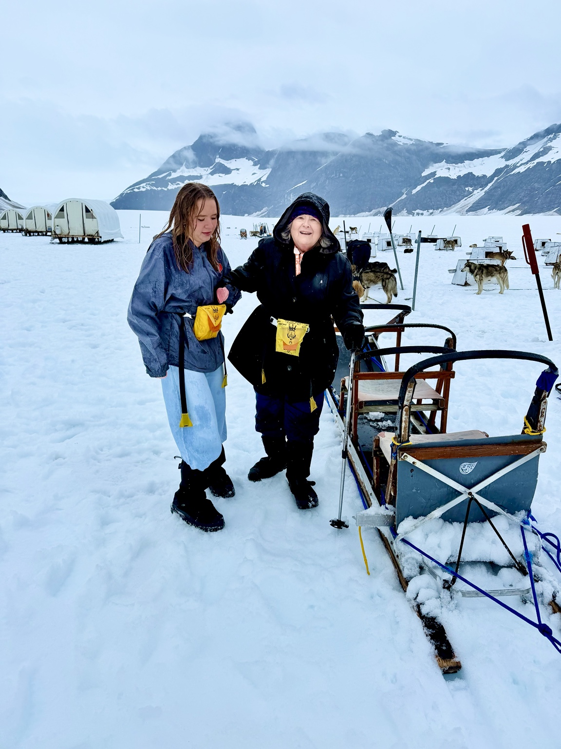 A younger family member steadying my mother as she walks across the snow toward a dog sled