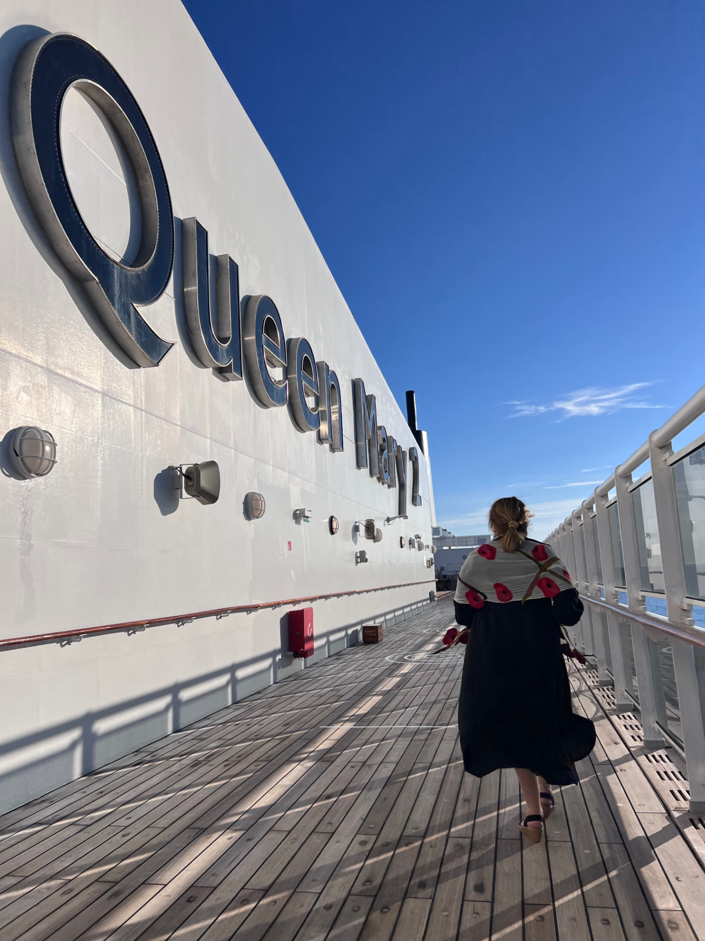 A woman walking along the teak promenade deck beneath the Queen Mary 2 name on the ship's hull