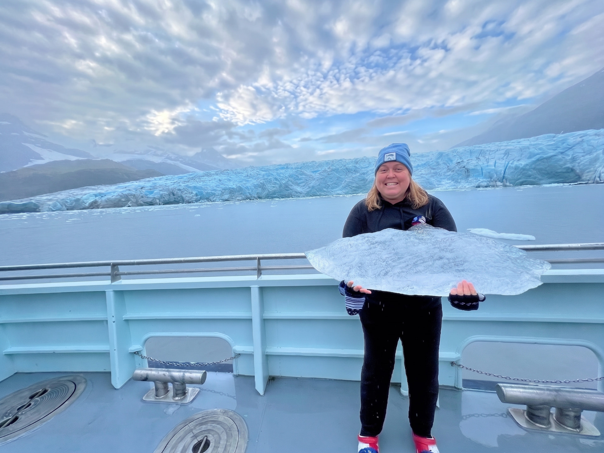 Christine holding a large piece of glacier ice on a ship deck with a blue glacier and mountains behind her in Alaska