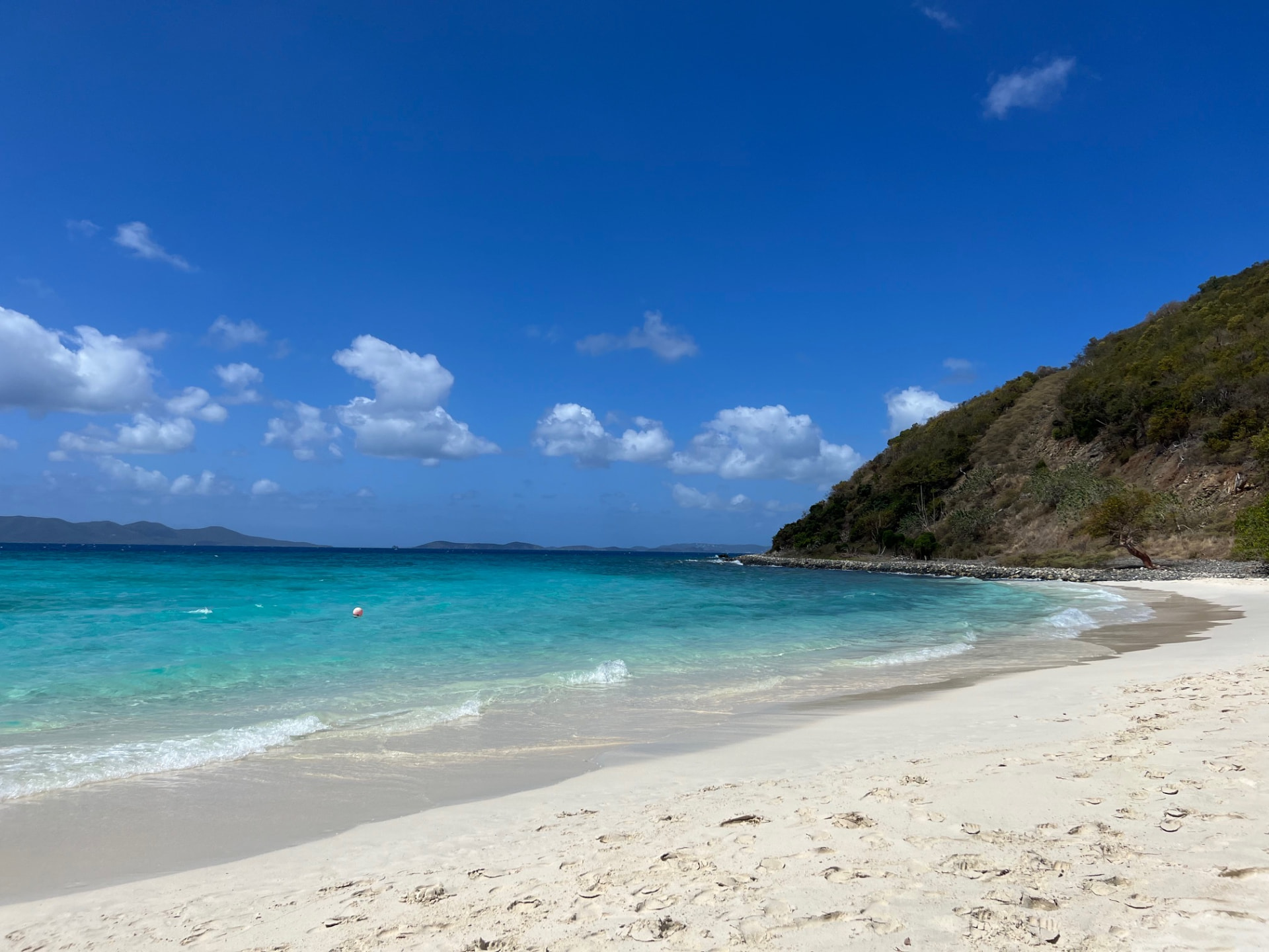 Empty white-sand Caribbean beach curving along turquoise water beneath a green hillside and bright blue sky