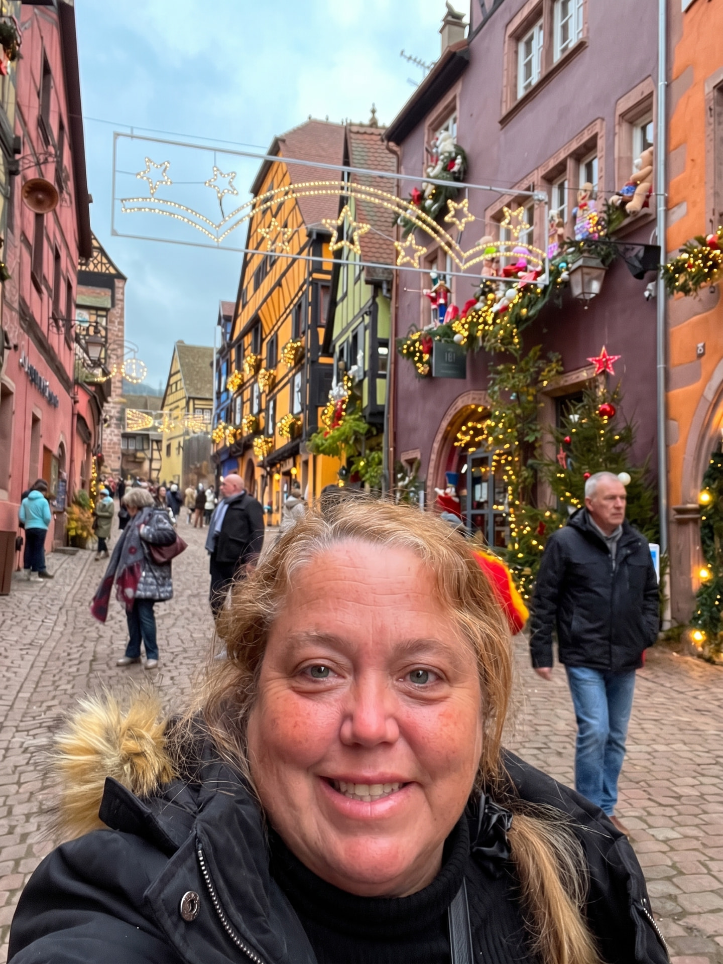Christine Scheeler smiling on a cobblestone street in Riquewihr, France, lined with half-timbered houses and Christmas lights
