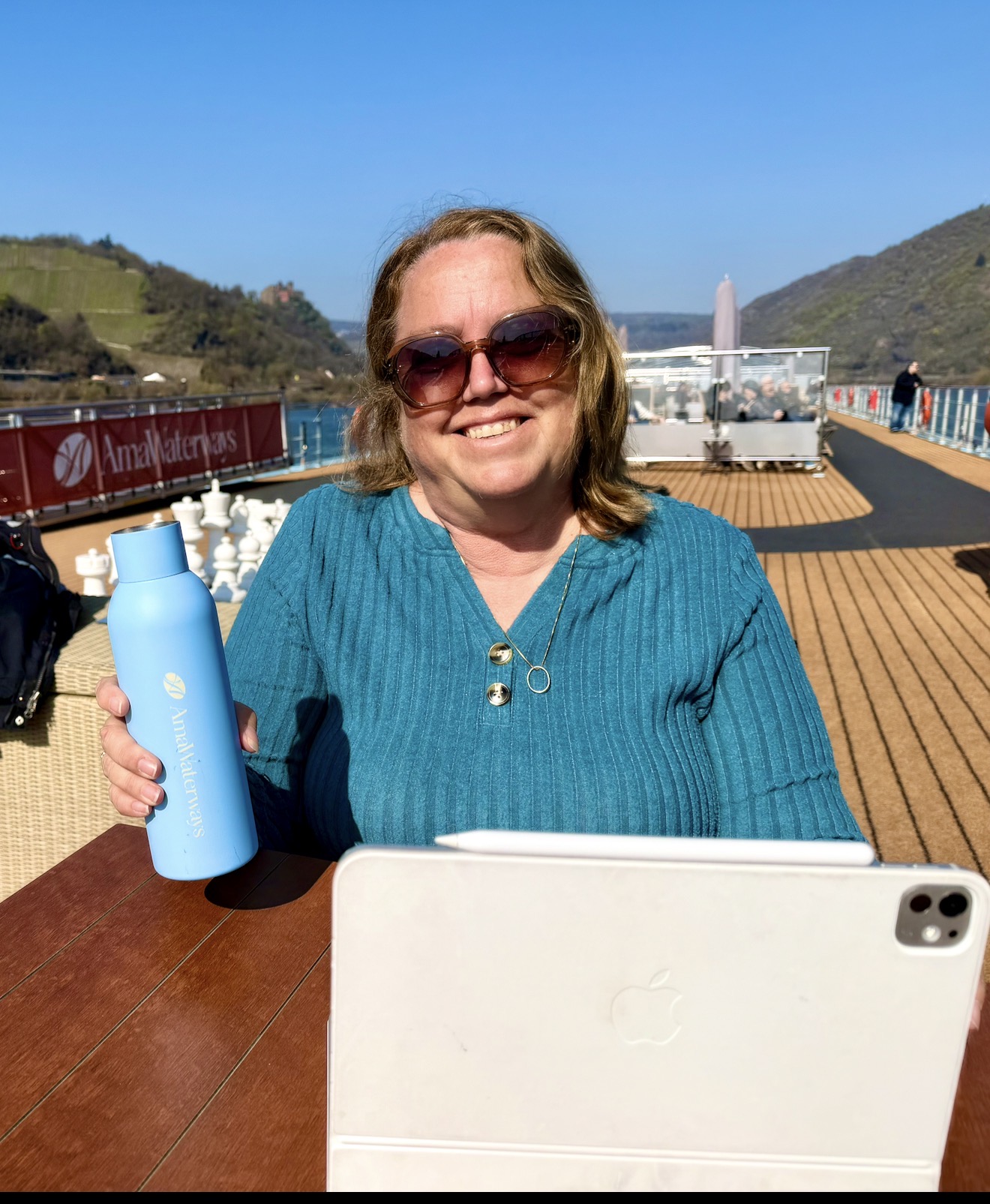 Christine Scheeler working on an iPad on the sun deck of an AmaWaterways river cruise ship, holding a branded water bottle with vineyard hills along the river behind her