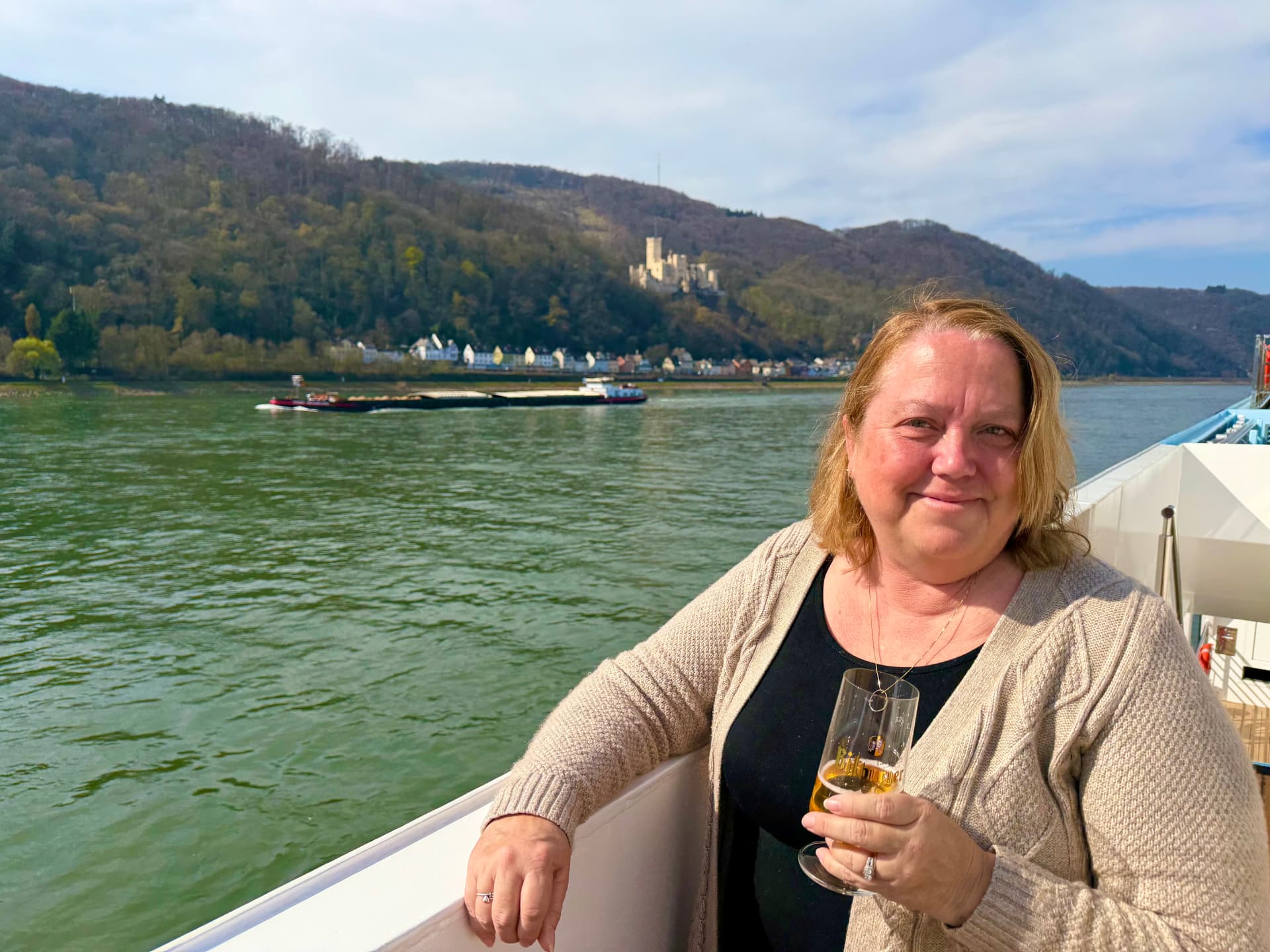 Christine Scheeler on a Rhine river cruise with a castle on the hillside, a barge on the water, and forested hills in the background